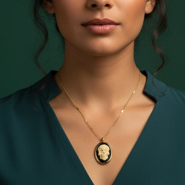 A gold skull pendant necklace on a gold chain, displayed on a brown cardboard tag from Amano Studio. The oval pendant features a gold skull on a black background, with small dried flowers placed on either side of the tag on a white wooden surface.